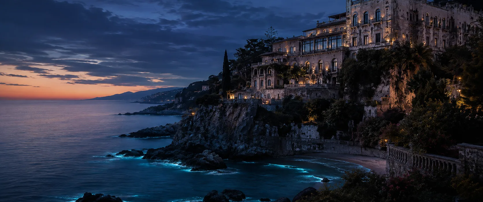A lit clifftop resort above a dark cove after sunset; the water along the rocks glows blue.